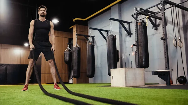 Full length of bearded sportsman holding battle ropes in gym - foto de stock