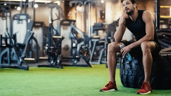 Tired sportsman in earphone resting on car tire and holding sports bottle in gym — Fotografia de Stock