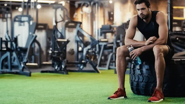 Tired sportsman in earphone listening music, sitting on car tire and puffing cheeks in gym — Foto stock
