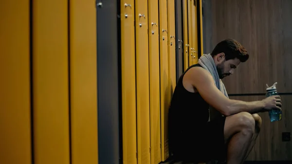 Side view of sportsman sitting on bench and holding sports bottle in locker room — стоковое фото