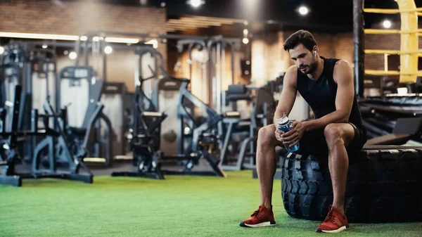 Exhausted sportsman in earphone sitting on car tire and holding sports bottle in gym — Stock Photo