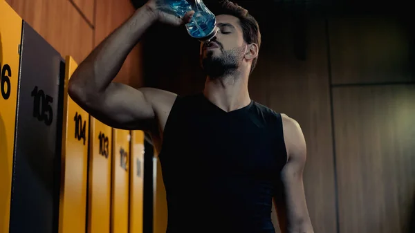 Bearded sportsman drinking water from sports bottle in locker room — Stockfoto