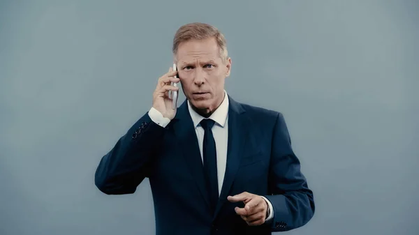 Homme d'affaires mature pointant du doigt et parlant sur smartphone isolé sur gris — Photo de stock