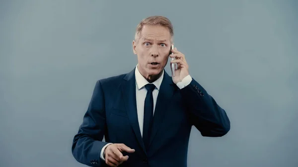 Шокований бізнесмен розмовляє на смартфоні і вказує пальцем ізольовано на сірому — стокове фото