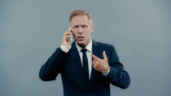 Mature businessman gesturing and talking on smartphone isolated on grey — Foto stock