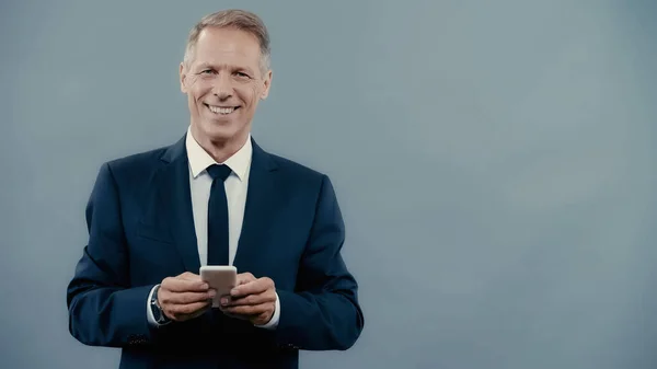 Cheerful mature businessman in suit holding smartphone and looking at camera isolated on grey — Stock Photo