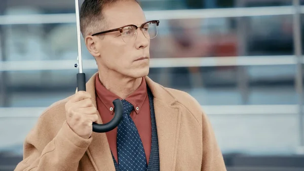 Mature businessman in eyeglasses holding umbrella on urban street — Stock Photo