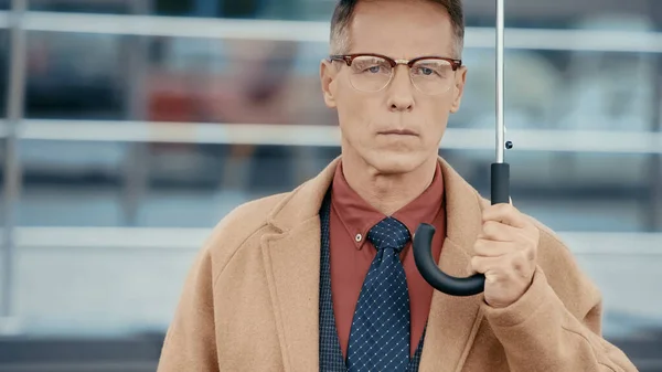 Middle aged businessman in coat holding umbrella and looking at camera on urban street — Stock Photo