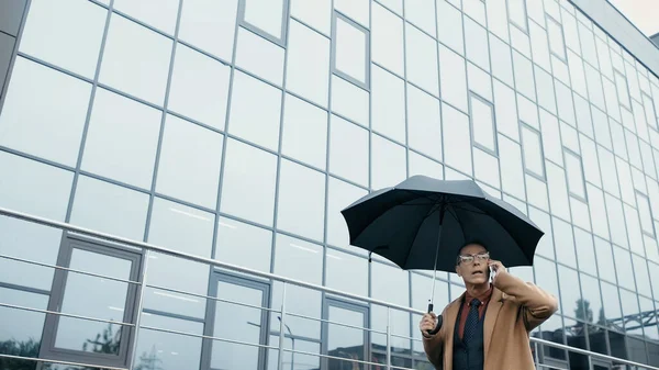 Businessman holding umbrella while talking on smartphone and walking near building outdoors — Fotografia de Stock