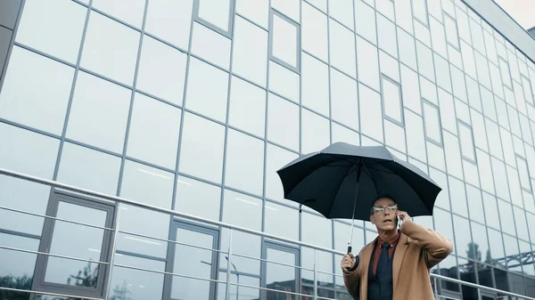 Businessman holding umbrella while talking on smartphone and walking near building outdoors — стоковое фото