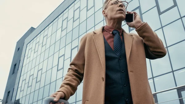 Low angle view of businessman talking on smartphone and holding suitcase on urban street — Foto stock