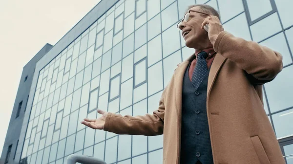 Low angle view of cheerful businessman in coat talking on smartphone on urban street — Stock Photo