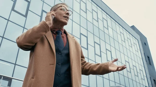 Low angle view of businessman in coat talking on cellphone near building on urban street — стоковое фото