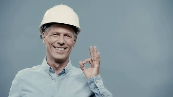 Smiling engineer in hardhat showing okay gesture isolated on grey — стоковое фото