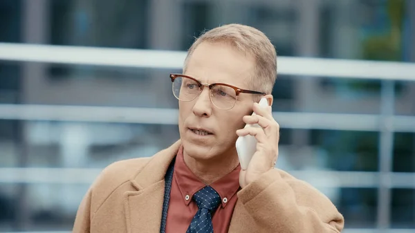 Middle aged businessman in eyeglasses and coat talking on cellphone outdoors — Stock Photo