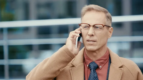 Mature businessman in coat talking on mobile phone on urban street — Stockfoto