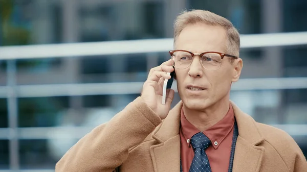 Mature businessman in coat talking on mobile phone on urban street — Stock Photo