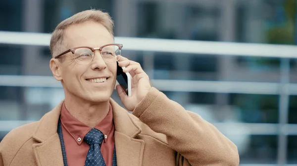 Happy mature businessman in coat talking on smartphone outdoors — Stock Photo