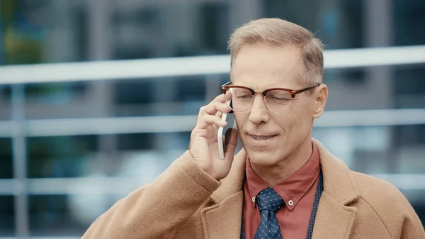 Homme d'affaires souriant en manteau parlant sur un téléphone portable dans une rue urbaine — Photo de stock