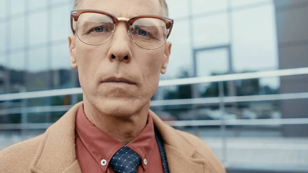 Middle aged businessman in eyeglasses and coat looking at camera on urban street — Stock Photo