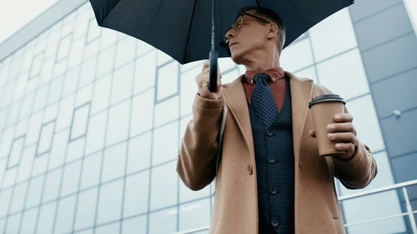 Low angle view of businessman holding coffee to go and umbrella on urban street — Stockfoto