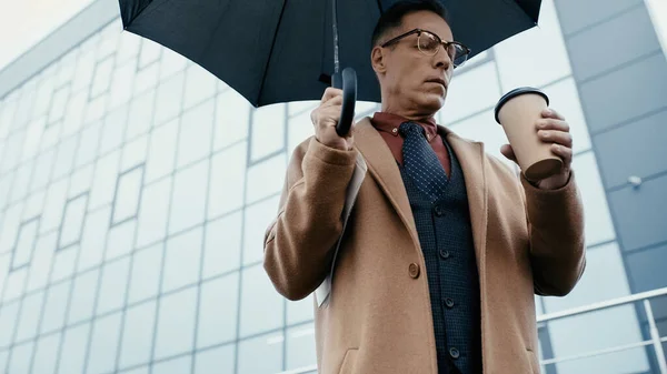 Низький кут зору бізнесмена в пальто тримає паперовий стаканчик і парасольку на міській вулиці — стокове фото