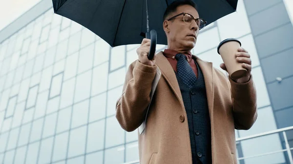 Low angle view of businessman in coat holding paper cup and umbrella on urban street — Stock Photo