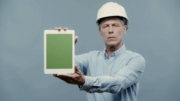 Blurred engineer in helmet showing digital tablet with green screen isolated on grey — Stock Photo