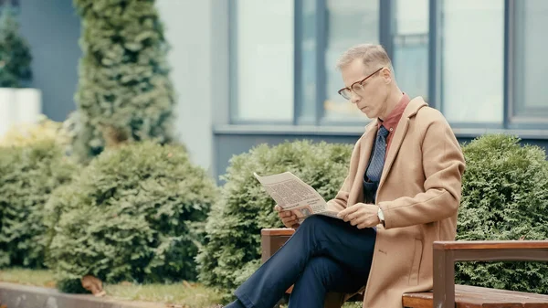 Mature businessman in coat reading newspaper on bench on urban street - foto de stock
