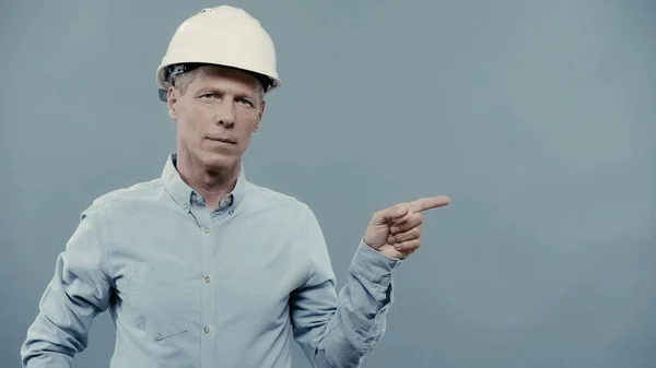 Mature engineer in helmet pointing with finger and looking at camera isolated on grey — Stockfoto