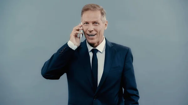 Positive businessman in formal wear talking on smartphone isolated on grey — Stock Photo