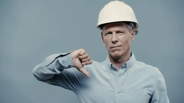 Middle aged engineer in hardhat showing dislike sign isolated on grey — стоковое фото