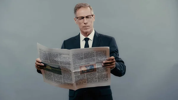 Middle aged businessman in suit reading newspaper isolated on grey — стоковое фото