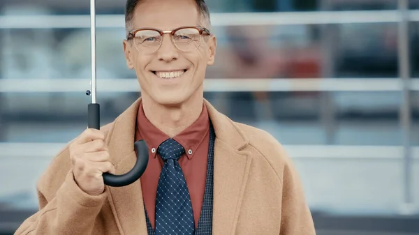 Smiling businessman in coat and eyeglasses holding umbrella on urban street — Photo de stock