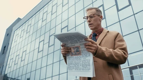 Low angle view of businessman in coat and earphone reading newspaper on urban street — Stock Photo