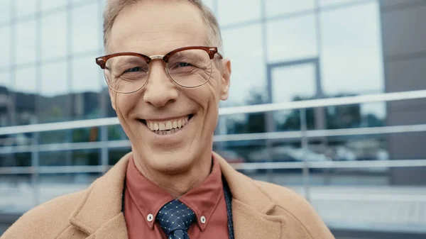 Portrait of happy businessman in coat and eyeglasses looking at camera outdoors — Stock Photo