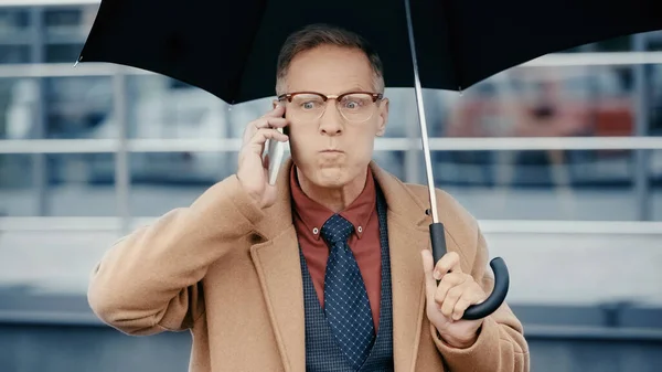 Homme d'affaires en colère parler sur smartphone et tenant parapluie à l'extérieur — Photo de stock