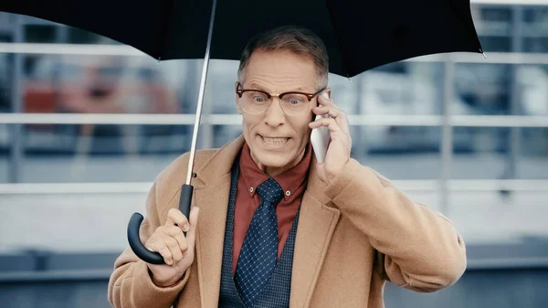 Aggressive businessman in coat talking on smartphone and holding umbrella outdoors — Stock Photo