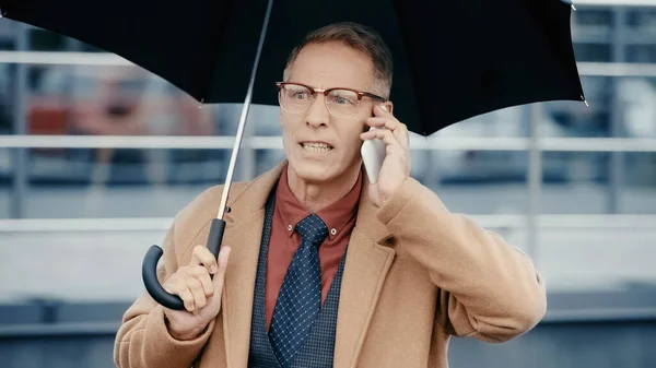 Irritated businessman in coat talking on smartphone and holding umbrella outdoors — Stock Photo