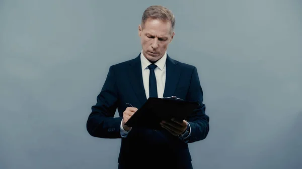 Empresario en traje escribiendo en portapapeles aislado en gris - foto de stock