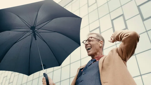 Low angle view of positive businessman in earphone holding umbrella on urban street — Stock Photo