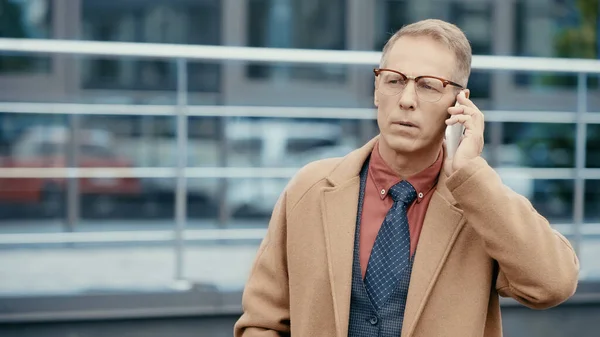 Mature businessman in eyeglasses and coat talking on smartphone on urban street — Stockfoto