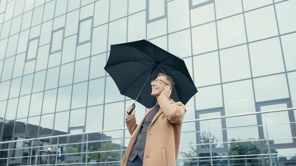 Smiling businessman in coat holding umbrella near building outdoors — Foto stock