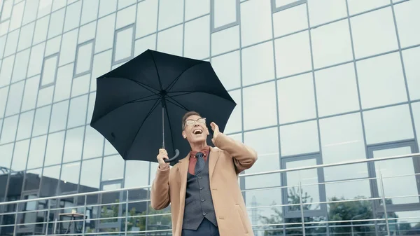 Happy businessman in coat and earphones holding umbrella on urban street — Stock Photo