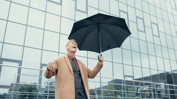 Positive businessman in coat using earphones and holding umbrella on urban street — Foto stock