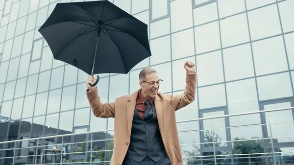 Cheerful businessman in earphone holding umbrella near building on urban street — стоковое фото