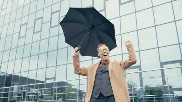 Excited businessman in coat using earphones and holding umbrella on urban street — Fotografia de Stock