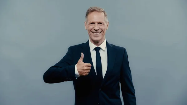 Smiling businessman showing thumb up and looking at camera isolated on grey — Stock Photo