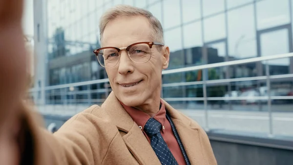 Smiling businessman in coat and eyeglasses looking at camera on urban street — Stock Photo