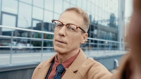 Middle aged businessman in eyeglasses and coat looking at camera outdoors — Fotografia de Stock